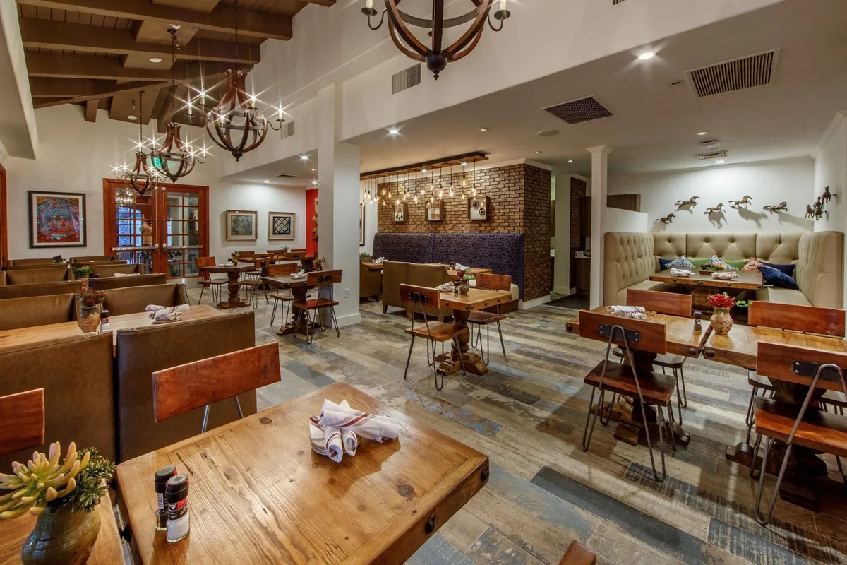 A warmly lit interior dining space featuring wooden tables, plush seating with cushions, elegant chandeliers, and vibrant artwork on exposed brick walls. The space is inviting and captures the atmosphere of a place where guests enjoy meals and conversation.