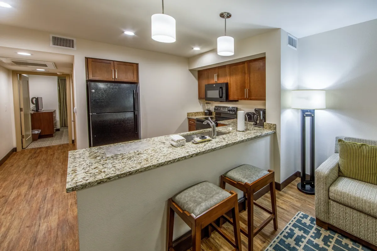 An open-concept living area with a modern kitchen featuring granite countertops, wooden cabinets, and bar stools, seamlessly connecting to a cozy sofa. The space is designed for cooking, entertaining, and relaxing, offering both functionality and warmth.