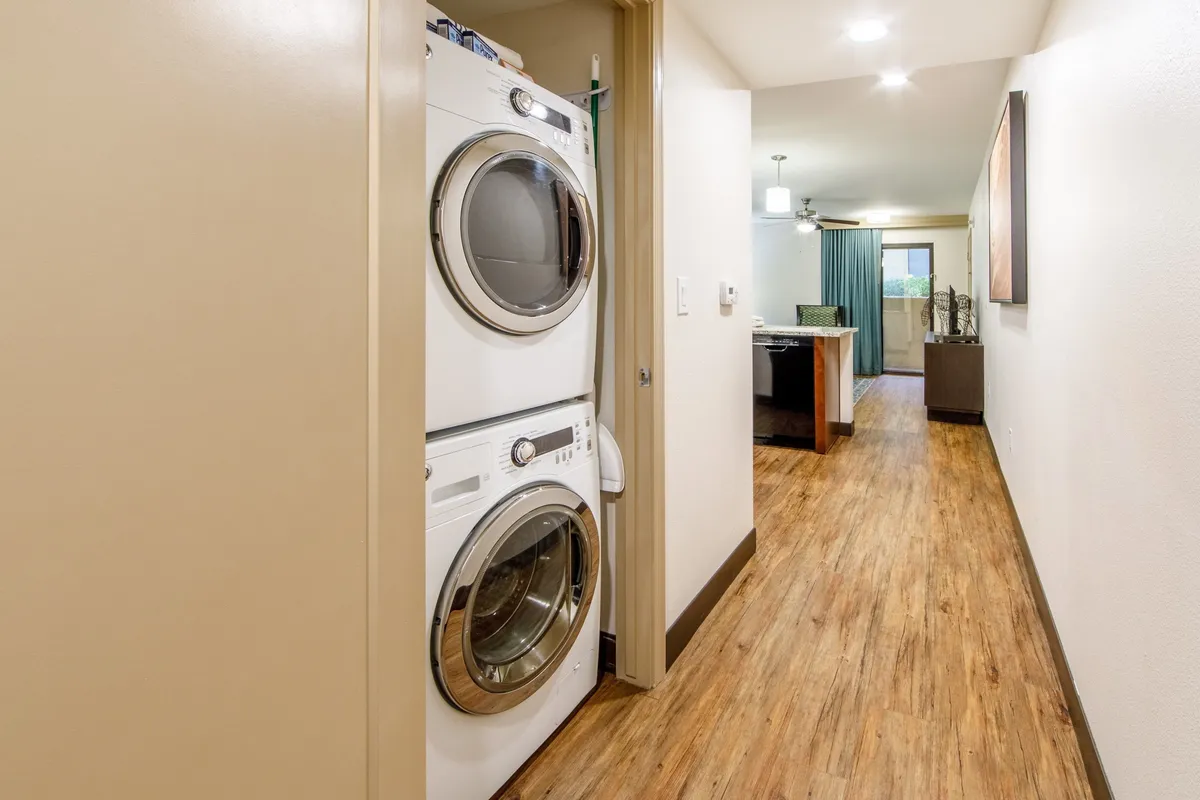 A compact yet functional laundry area featuring a stacked washer and dryer, wooden flooring, and a view into the cozy living area, emphasizing home-like convenience within the villa.