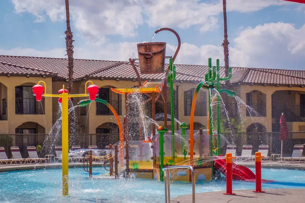 A colorful water play structure set in a large pool area surrounded by palm trees and plenty of lounge chairs. Children are engaged in playful splashing while families gather nearby, all bathed in warm sunlight.