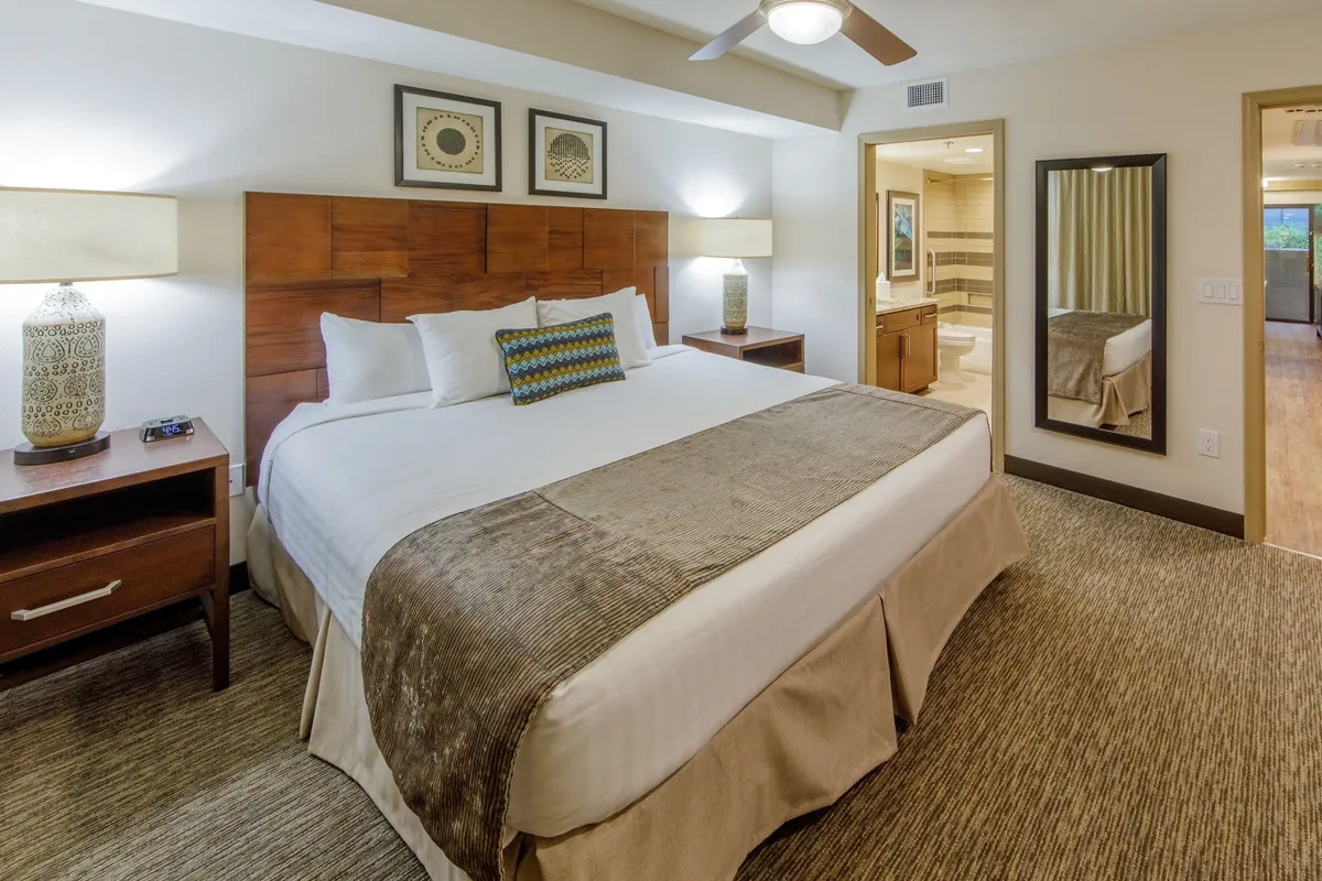 A serene and inviting bedroom decorated with soft, neutral tones. The space features a comfortable bed with decorative pillows, matching nightstands with elegant lamps, and a large mirror that reflects natural light streaming in through a nearby window. The atmosphere is tranquil and designed for relaxation, with subtle contemporary decor touches.