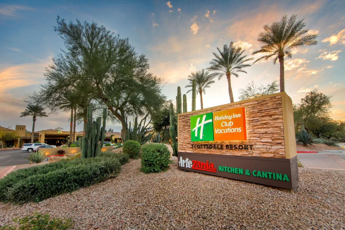 A classic Holiday Inn sign anchors a beautifully landscaped desert setting, framed by palm trees and local flora. The image captures the resort’s exterior as guests might see it when exploring the property or heading out to nearby attractions such as golf courses.