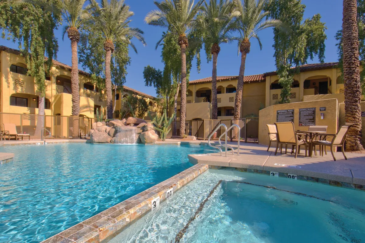 An inviting resort-style swimming pool is surrounded by palm trees and plush lounge chairs, with a gentle waterfall feature creating a relaxing ambiance. The scene is bathed in warm light, perfect for swimming, sunbathing, or unwinding by the pool after a day of activity.