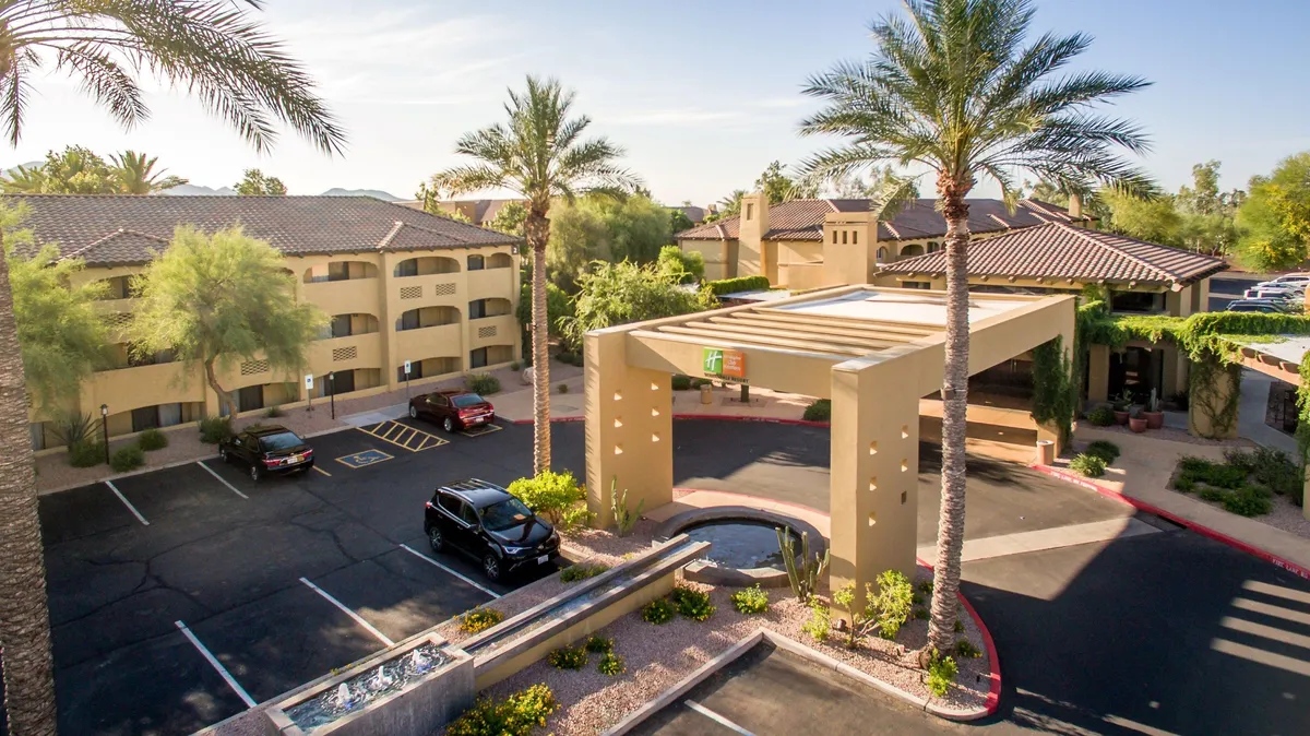 This image captures the inviting exterior of the property, featuring palm trees, a welcoming entryway structure, a central fountain, and the parking area. The scene hints at the resort's accessibility and sets the tone for family arrivals, with visible landscaping and social spaces.
