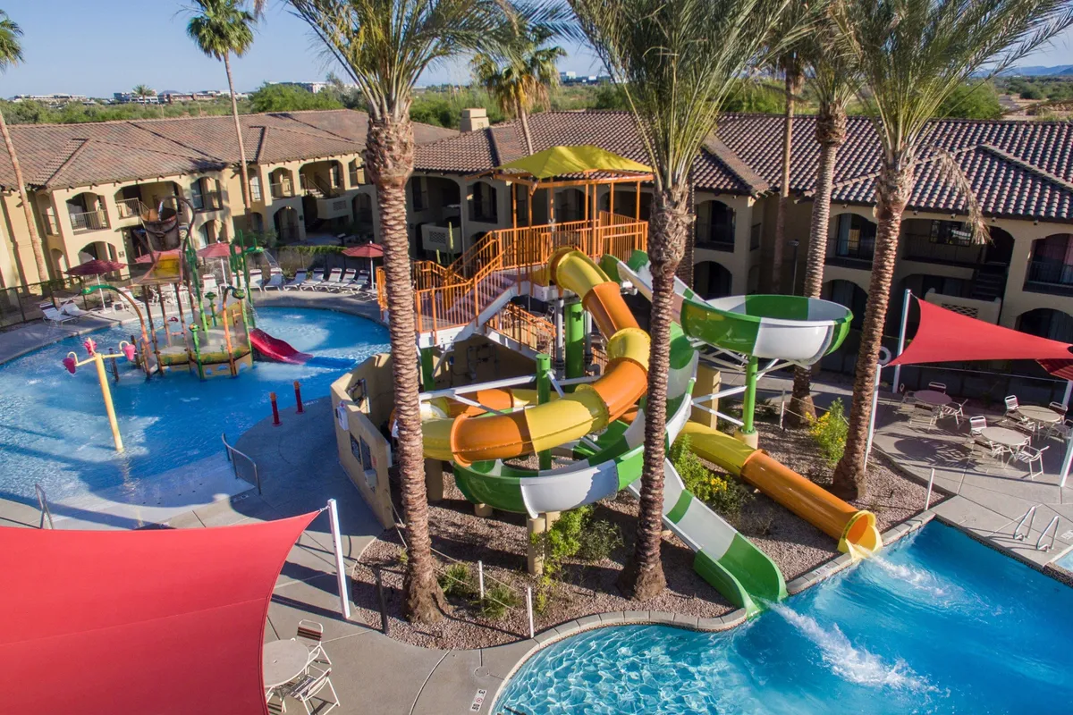 A vibrant outdoor pool scene featuring twisting water slides, a spacious swimming pool, palm trees, and a children’s play area. Families and kids enjoy lively water play under the sun, with lounge chairs and gathering spaces visible nearby.