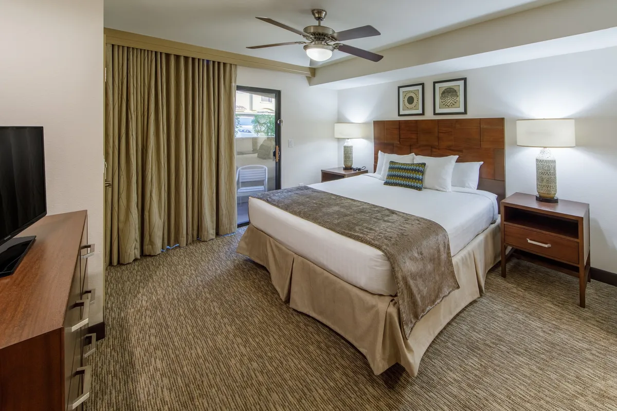 A tranquil king bedroom featuring a plush king-size bed adorned with crisp linens, flanked by nightstands and soft drapes. A television is mounted opposite the bed, and a door to a patio lets in natural light, enhancing the room's restful ambiance.
